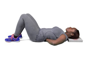 A person lying on their back with a pillow under the lower back, wearing a sports bra and leggings, resting with one foot in the air, with a blue sneaker visible.