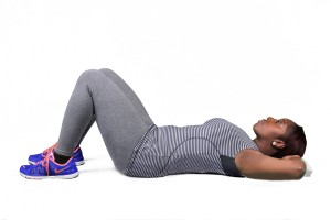 Woman in a gym, stretching on the floor with her arms behind her head.