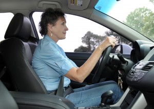The image is a photograph of an older woman driving a car with her hands on the steering wheel, wearing a blue shirt and seated in a vehicle s driver s seat.