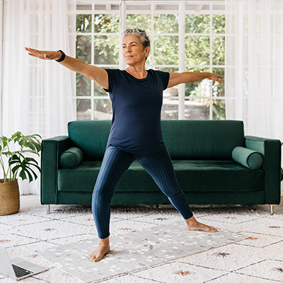 The image shows an older woman in a dark blue tank top and light blue leggings, stretching her arms upwards while standing on a wooden floor. She is positioned in front of a couch with a white cushion, which has a green throw blanket draped over it. A potted plant is visible to the left of the couch. The room appears to be a living area, and there s a laptop on a coffee table in the foreground.