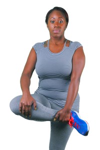 A woman in a striped tank top and leggings is standing with one hand on her knee, wearing a black headband and holding a blue shoe in her other hand.