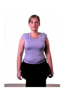 The image shows a woman standing with her hands on her hips, wearing a sleeveless top and black pants. She appears to be in a gym or exercise setting, as suggested by the presence of what looks like an exercise machine or equipment behind her.