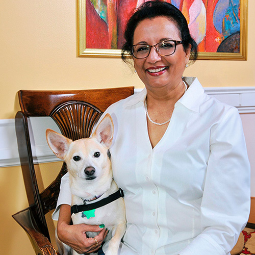 The image shows a woman holding a small dog, both seated in front of a painting on the wall.