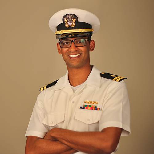 The image shows a person in military attire, specifically an officer s uniform with rank insignia and a cap, standing confidently with arms crossed.