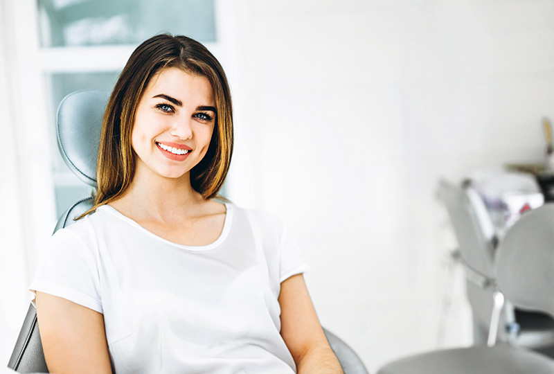 A woman is seated in a dental office chair, smiling at the camera.