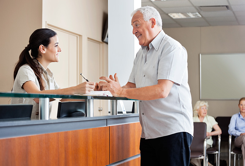 The image depicts an older man standing at a counter, engaged in conversation with a woman who is seated behind the counter.