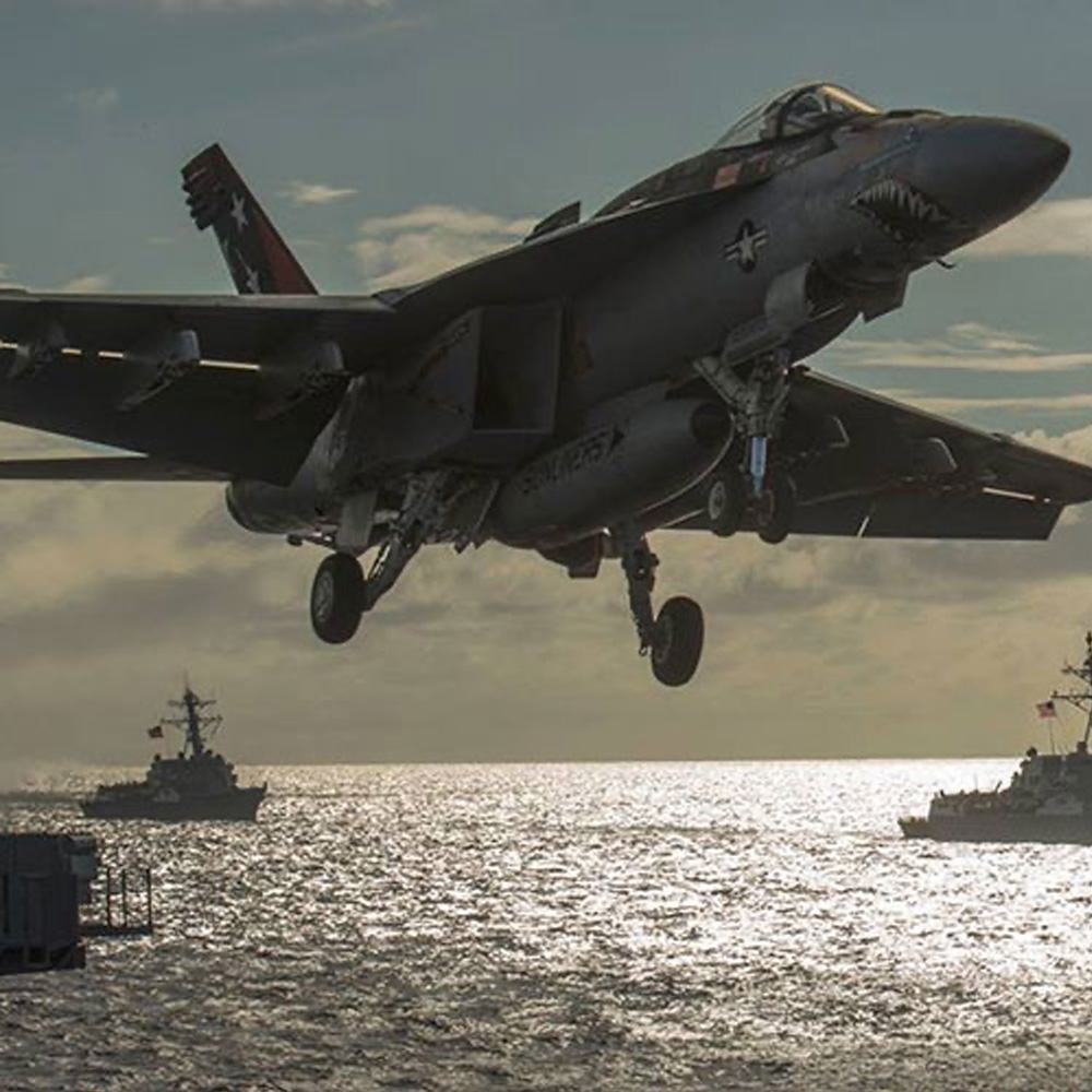 The image shows a fighter jet flying low over the ocean, with a military vessel and a dock in the background.