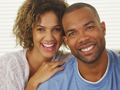 A man and a woman are smiling at the camera, with the man wearing a short beard and both dressed in casual attire.